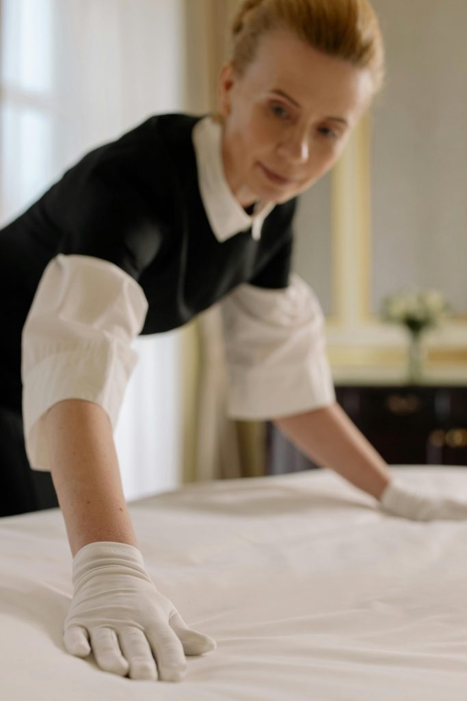 A housekeeper elegantly arranges bed sheets in a luxurious hotel room, showcasing hospitality expertise.